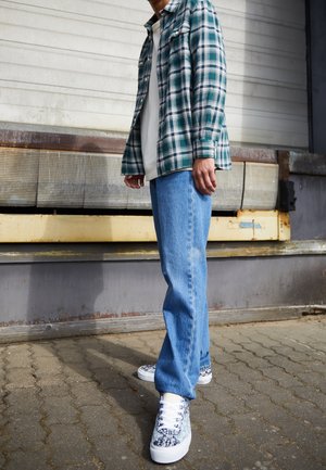 Personne portant un jean bleu, des baskets bleues et blanches à motifs, un T-shirt blanc et une chemise à carreaux verte, debout sur un sol pavé près d'un mur industriel.