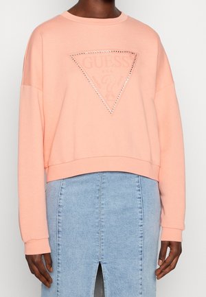 Kvinde iført en ferskenfarvet langærmet sweatshirt med et nittet Guess-logo trekant og en lyseblå denim nederdel med frontspalte.