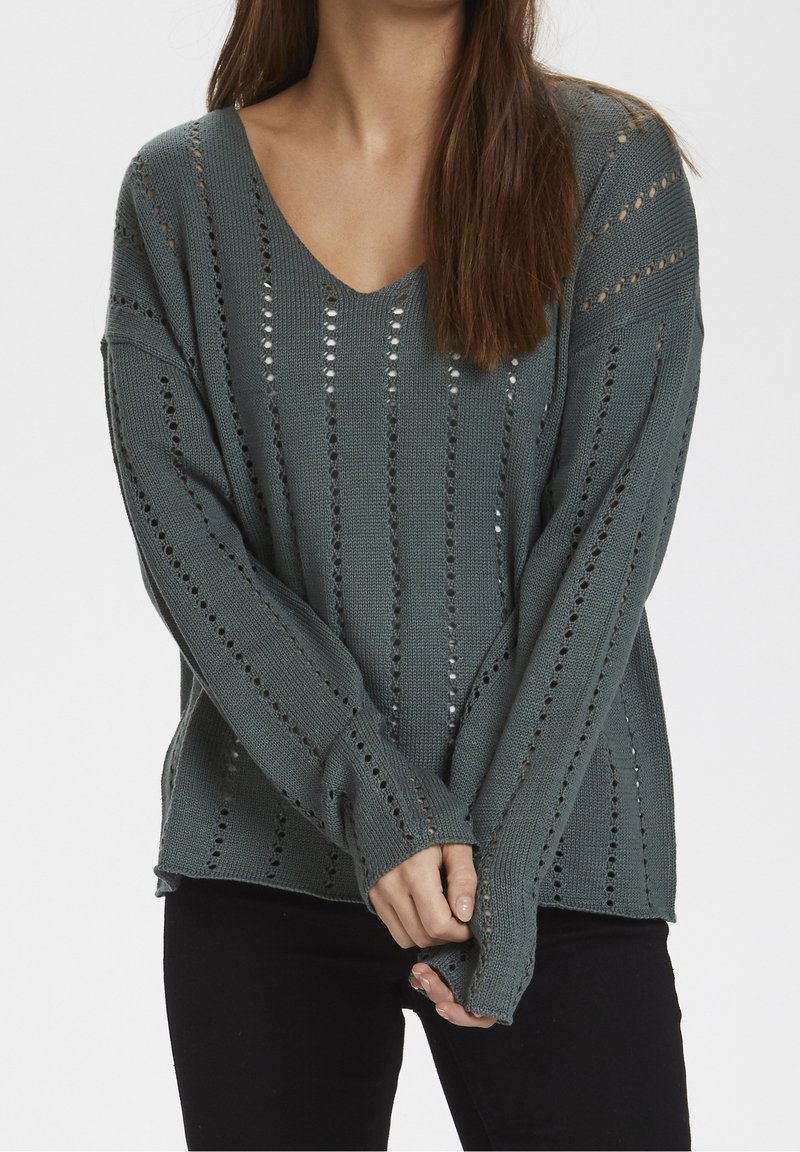 Femme portant un pull ample en tricot gris charbon avec des motifs ajourés verticaux et un pantalon noir, debout les mains croisées.