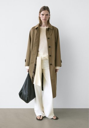 Vrouw staand, gekleed in een bruine trenchcoat, crèmekleurige trui, witte wijd uitlopende broek, zwarte sandalen en met een grote zwarte handtas in haar hand.