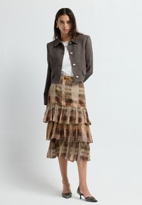 Bruine gestructureerde cropped jas met gouden knopen, gecombineerd met een gelaagde, gemusterde midi-rok in aardetinten en spitse zwarte hakken.