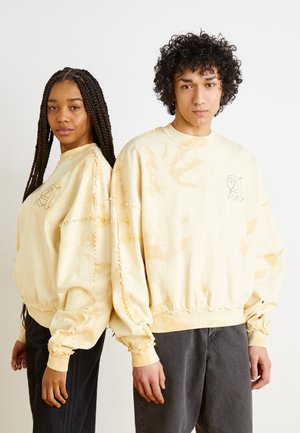 Sweatshirts jaune tie-dye avec des poignets et un ourlet élastiques, arborant un petit logo brodé. Les modèles portent des jeans foncés et affichent des profils latéraux.