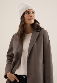 Grijze wolmix jas met een geknipt kraag, twee knopen en zijzakken, gedragen over een witte top en gecombineerd met een licht gestreepte gebreide beanie.
