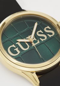 Złoty zegarek z zieloną teksturowaną tarczą, z wyraźnym logo "GUESS", białymi wskazówkami godzinową i minutową, umieszczony na czarnym pasku.
