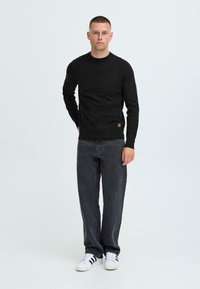 Zwarte lange mouwen trui van gebreide stof, gecombineerd met donkere jeans en witte sneakers, met een klein merklabel aan de onderkant.