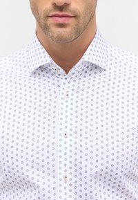 Camicia bianca con motivo floreale in blu e grigio, dotata di colletto a punta, pattina con bottoni e texture liscia.