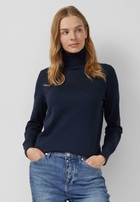 Femme aux cheveux blonds portant un pull à col roulé bleu marine et un jean bleu, les mains dans les poches, se tenant devant un fond uni.