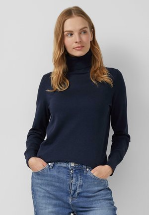 Femme aux cheveux blonds portant un pull à col roulé bleu marine et un jean bleu, les mains dans les poches, se tenant devant un fond uni.