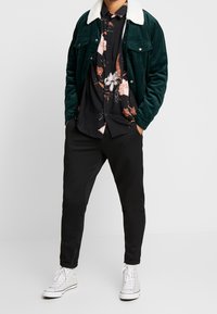 Veste en velours côtelé vert avec un col en fausse fourrure blanche portée sur une chemise noire à fleurs, associée à un pantalon de survêtement noir et des baskets blanches.