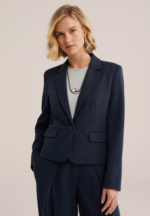 Donna bionda che indossa un blazer blu navy e pantaloni abbinati, top grigio, collana e orecchini argentati, in piedi con una mano in tasca.