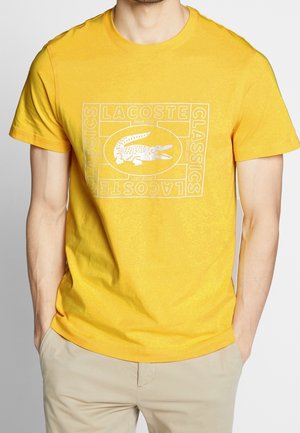 Mand iført en knaldgul Lacoste t-shirt med hvid krokodille-logo og tekst, kombineret med beige bukser, hænder i lommerne.
