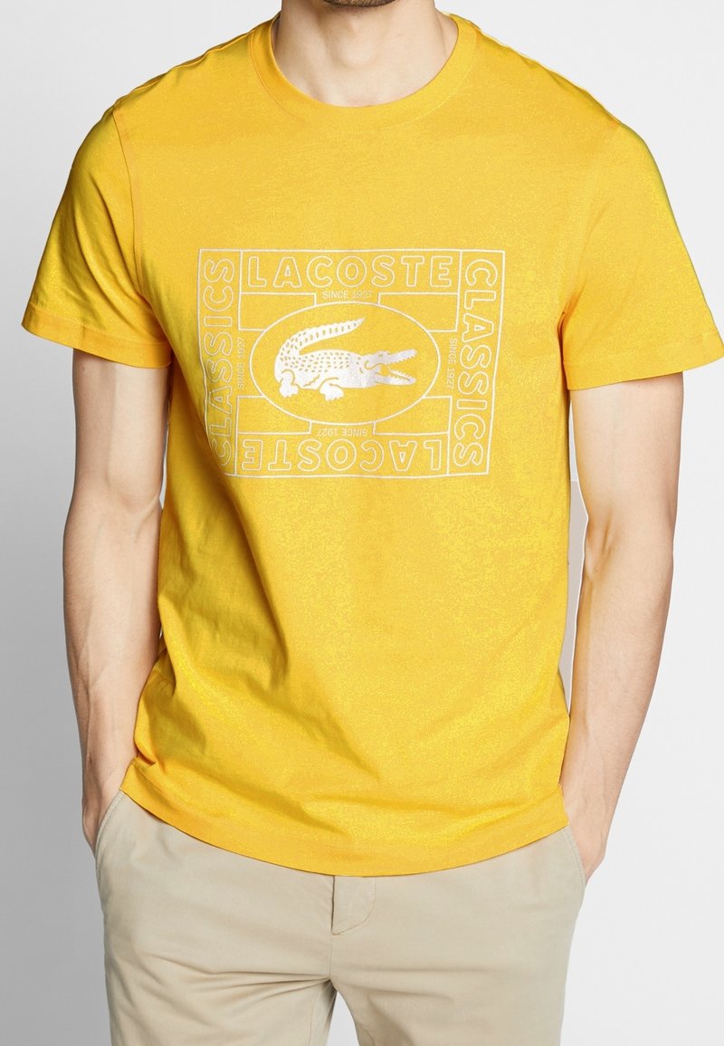 Homme portant un t-shirt Lacoste jaune vif avec un logo crocodile blanc et un texte, associé à un pantalon beige, mains dans les poches.