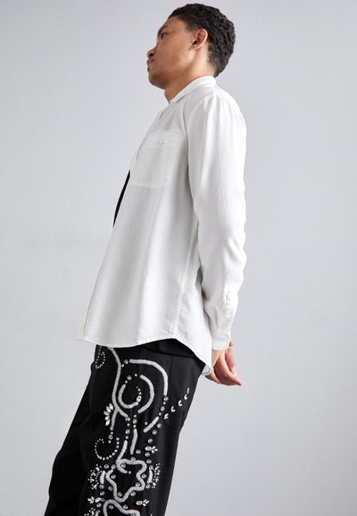 Hosszú ujjú, fehér ing ívelt aljjal, fekete nadrággal párosítva, amely bonyolult ezüst díszítésekkel és hímzett mintákkal rendelkezik.