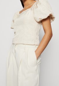 Haut beige clair à manches courtes avec un décolleté carré et des manches bouffantes, fabriqué à partir d'un tissu texturé. Associé à un pantalon crème taille haute.