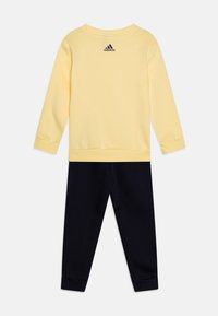 Sudadera amarilla de manga larga y cuello redondo, combinada con pantalones azul marino. Logo de Adidas en negro en la parte posterior de la sudadera.