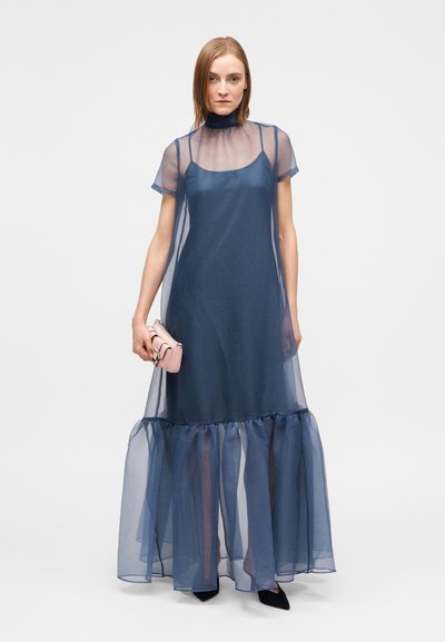 Γυναίκα με διαφανές μάξι φόρεμα σε ναυτικό μπλε χρώμα με βολάν στο τελείωμα, κρατώντας μικρό τσαντάκι σε ανοιχτό ροζ, φορώντας μαύρες μυτερές γόβες, στέκεται μπροστά σε λευκό φόντο.