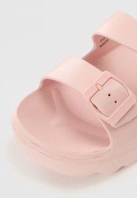 Rosa gummisandal med två breda remmar, en rektangulär spänne och en texturerad sula för grepp. Slät yta med små tonvariationer.