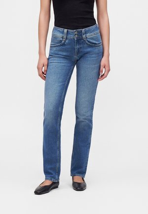 Femme portant un jean bleu taille haute à jambes droites avec fermeture à double bouton, associé à un haut noir sans manches et des ballerines noires.