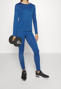 Top atletico blu a maniche lunghe e leggings abbinati in un tessuto testurizzato, abbinati a scarpe da ginnastica nere e in procinto di sollevare dei manubri.