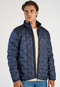 Marinblå puffjacka med strukturerad yta, hög krage och mörka axlar. Lättviktig, med frontdragkedja och sidofickor.