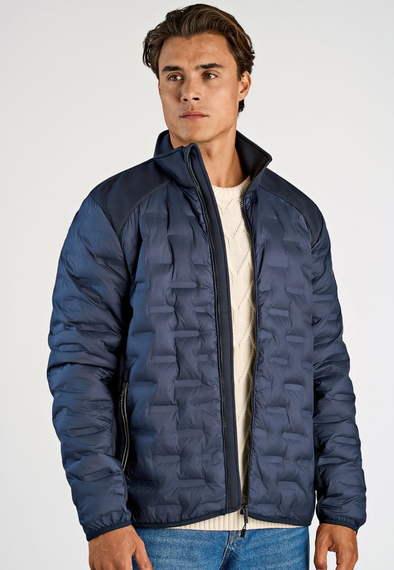 Marinblå puffjacka med strukturerad yta, hög krage och mörka axlar. Lättviktig, med frontdragkedja och sidofickor.