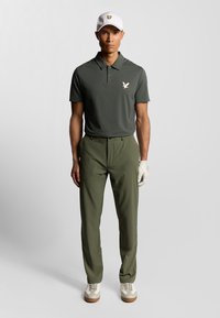 Polo shirt verde scuro con un piccolo logo chiaro, abbinato a pantaloni verde oliva. Un cappellino bianco con logo giallo e scarpe bianche completano il look.