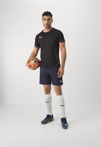 Camisola atlética preta, calções azuis, meias brancas e botas de futebol azuis com detalhes verdes. A segurar uma bola de futebol laranja e preta.