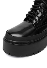 Cizme negre lucioase până la gleznă, cu vârf rotund, talpă groasă și profil texturat din cauciuc. Dispune de șireturi negre și găuri pentru șireturi.