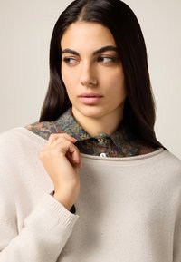 Beige Pullover mit Rundhalsausschnitt und feiner Strickstruktur, über einem dunkelblauen Hemd mit floralem Muster und Knöpfen getragen.