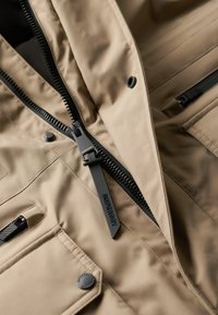 Chaqueta beige con un tejido texturizado, cremalleras negras, una pestaña de marca y múltiples bolsillos con botones a presión.
