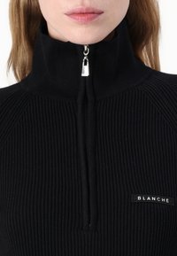Pulover negru cu fantele, cu guler înalt, având un fermoar argintiu și o etichetă rectangulară cu textul "BLANCHE" pe partea stângă.