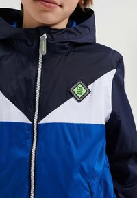 Huppari, jossa on vetoketju, sinivalkoisen ja sinisen paneelin yhdistelmä. Valmistettu sileästä kankaasta, jossa on brändilogo rinnassa ja vetoketjukiinnitys.