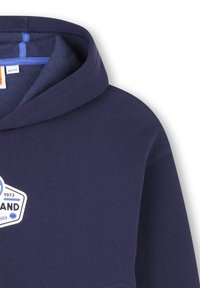 Felpa blu navy con cappuccio, caratterizzata da un patch logo bianco e blu. Realizzata in un tessuto morbido con una texture liscia e un design semplice.