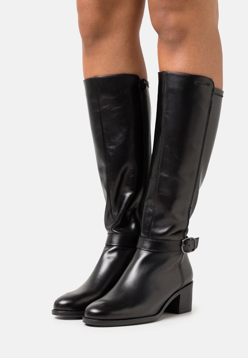 Bottes en cuir noires montantes jusqu'aux genoux, avec des talons blocs bas, des fermetures éclair latérales et des sangles décoratives à la cheville, portées sur des jambes nues devant un fond uni.
