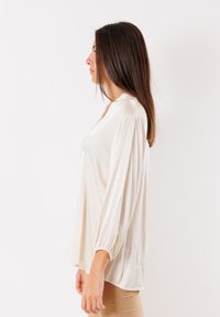 Blusa color crema con una texture morbida e setosa, caratterizzata da una vestibilità rilassata, maniche a pallone plissettate e un drappeggio sottile sull'orlo.