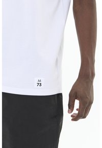 Fehér pamut póló, az alján egy kis textilcímkével, rajta "M 73", fekete sorttal párosítva. Simább textúra, laza fazon.