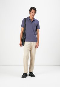 Jeune homme portant un polo violet, un pantalon beige, des chaussures noires et un sac à bandoulière noir, se tenant devant un fond blanc uni.