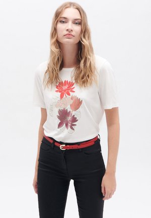 T-shirt blanc à manches courtes avec un motif floral dans des nuances de rouge et de rose, associé à un jean noir et une ceinture rouge avec une boucle en or.