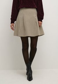 Jupe plissée à carreaux beige et noir, avec une taille haute et tombant mi-thigh, associée à des bottines noires et des collants.