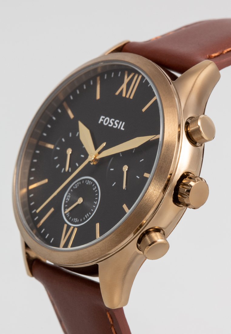 Reloj Fossil redondo con esfera negra, acentos dorados y marcadores en números romanos. Correa de cuero marrón, caja de acero inoxidable y esferas de cronógrafo.
