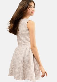 Teenager Mädchen mit langen braunen Haaren, das ein ärmelloses, hellfarbiges kariertes Kleid trägt, teilweise abgewandt ist und über ihre Schulter blickt.