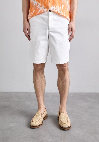 Weißes Baumwollshorts mit einem lässigen Schnitt, kombiniert mit hellbraunen geflochtenen Slippern mit einem kontrastierenden Rand. Der Hintergrund ist neutral.