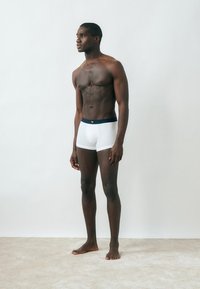 Αθλητικός άνδρας με σκούρο δέρμα, όρθιος ξυπόλυτος πάνω σε χαλί, φοράει λευκά μποξεράκια με σκούρο μπλε ζωνάκι και κοιτάει πλάγια με φόντο λευκό και απλό.