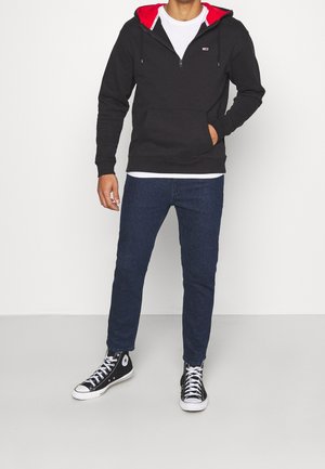 Sudadera negra con capucha forrada en rojo, bolsillo frontal y cierre de cremallera, combinada con vaqueros azul oscuro y zapatillas negras de caña alta.