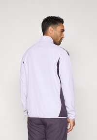 Jasnofioletowa sportowa kurtka z wysokim kołnierzem, ciemnoszarymi bocznymi panelami i gładką fakturą. Charakteryzuje się wzmocnionym przeszyciem i dopasowanym krojem.