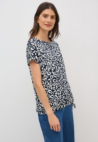 Marinblus med kort ärm och vitt leopardmönster. Lättviktsmaterial, rund halsringning, justerbar knytning i nederkant. Matchad med blå denimjeans.