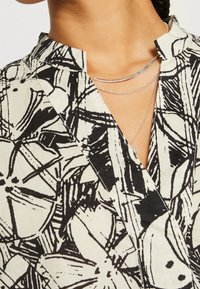 Gros plan d'une personne portant une chemise à motif floral abstrait noir et blanc avec col et colliers en argent superposés.