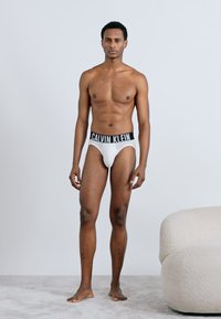 Λευκά ανδρικά σλιπάκια με μαύρη ζώνη "CALVIN KLEIN", παρουσιασμένα σε άντρα μοντέλο, επιδεικνύοντας εφαρμογή που τονίζει τη σιλουέτα και λείο ύφασμα.