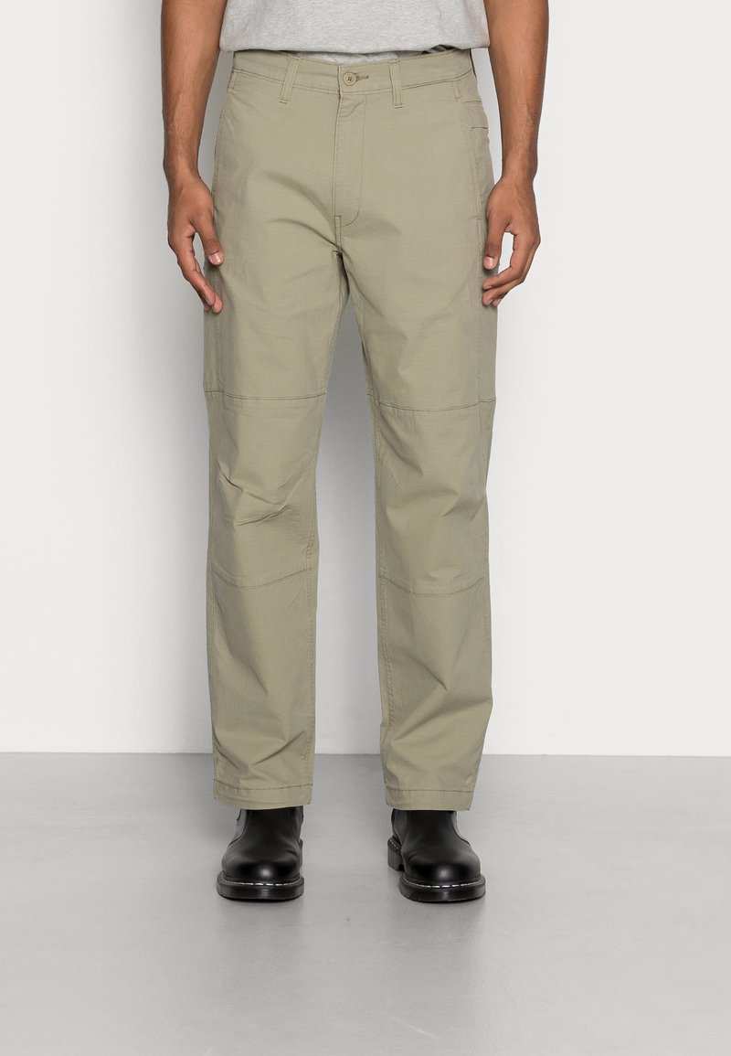 Homme portant un pantalon droit kaki clair et des bottines noires, debout contre un mur blanc uni et un sol gris.