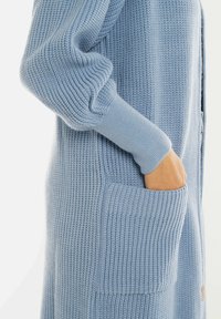 Hand in de zak van een lichtblauwe chunky gebreide cardigan met lange poffende mouwen en geribde manchetten.